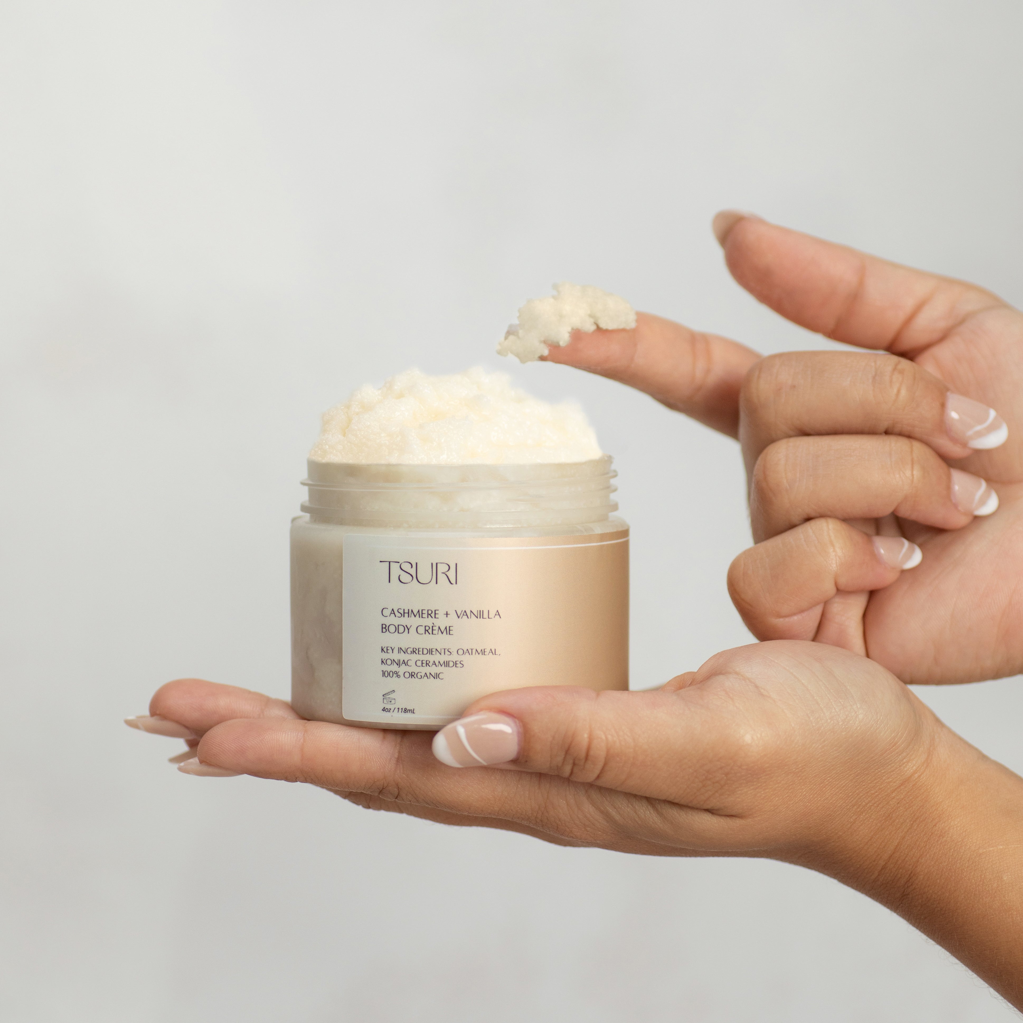 Person holding Tsuri Cashmere + Vanilla Body Crème jar, creamy texture visible on fingers, promotes deep hydration and softness.