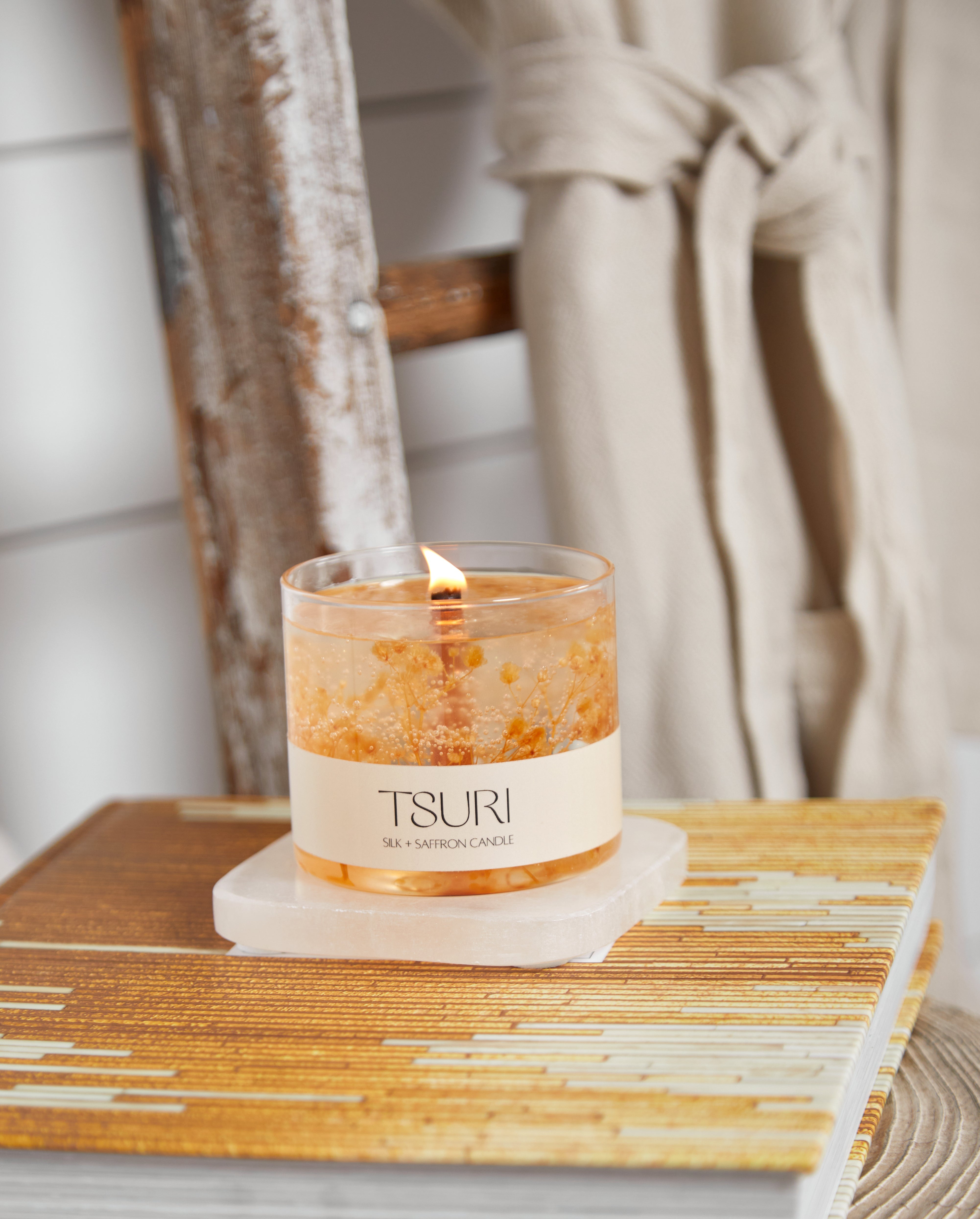Silk + Saffron Signature Scented Candle with wooden wick, burning on a book, next to a robe, creating a cozy ambiance.