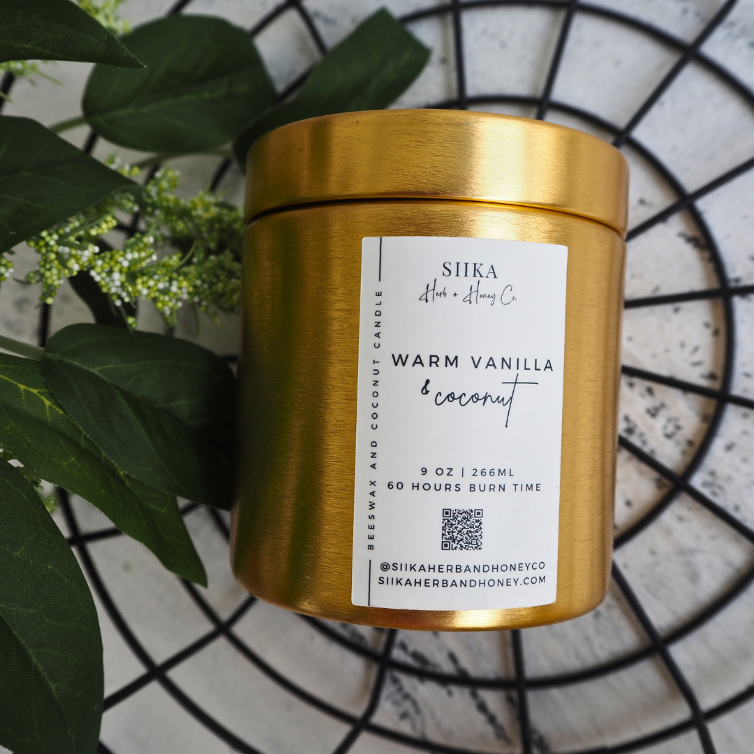 Alt Tag: Gold tin of SIIKA Warm Vanilla & Coconut beeswax candle, hand-poured for a clean burn, surrounded by leaves on tile background.
